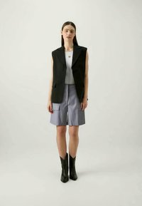 Black sleeveless blazer over a white tank top, paired with light blue cargo shorts and black ankle boots. Minimalistic design, structured shape.