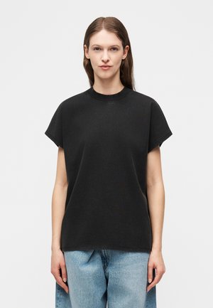 Γυναίκα με μακριά καστανά μαλλιά που φοράει μια άνετη μαύρη κοντομάνικη μπλούζα και ανοιχτό μπλε τζιν, στέκεται μπροστά σε ένα απλό λευκό φόντο.