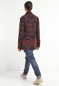 Femme aux cheveux bruns attachés, marchant, vêtue d'une veste à motifs rouge et noir, d'un jean bleu et de baskets beiges, sur un fond blanc.