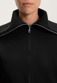 Musta takki, jossa on korkea kaulus, täysipitkä vetoketju ja teksturoitu kangas. Lähikuva keskittyy kauluksen ja vetoketjun yksityiskohtiin.