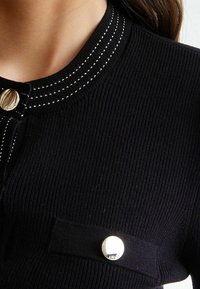 Sort ribstrikket top med rund halsudskæring og hvide syninger, lukning med guldknap og en påsyet lomme med guldknapdetalje.
