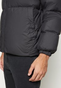 Veste matelassée noire avec poche latérale et poignet élastique sur la manche, portée avec un pantalon noir, mettant l'accent sur les détails du torse et des mains.