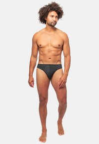 Schwarze Schwimmslips mit strukturiertem Muster, mit einem mittelhohen Schnitt und elastischem Bund, getragen von einem männlichen Model vor einem weißen Hintergrund.