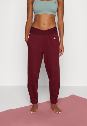 Kobieta stojąca boso na różowej macie do jogi, ubrana w zielony sportowy stanik i bordowe spodnie dresowe Adidas z logo na udzie.