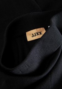 Crna dukserica s rebrastim ovratnikom, s bež etiketom na kojoj piše "JJXX" crnim slovima. Glatka tekstura; kratak pregled dekoltea.