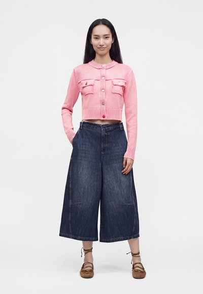 Jeune femme debout portant un cardigan court rose boutonné, des culottes en jean foncé à jambes larges et des chaussures plates marron à lacets sur un fond blanc uni.