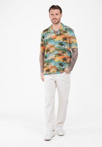 Homme tatoué portant une chemise à imprimé tropical coloré, un pantalon blanc et des baskets blanches, se tenant devant un fond blanc uni.