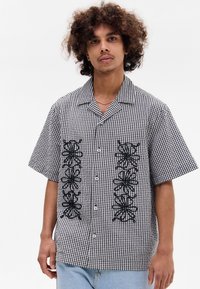 Camisa de manga curta com padrão de gingham em preto e branco, apresentando designs florais bordados a preto na parte da frente, e um fechamento de botões.
