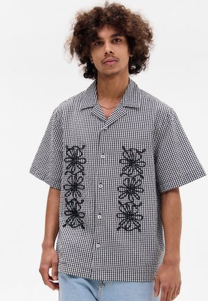 Camisa de manga corta con un patrón de cuadros en blanco y negro, que presenta diseños florales bordados en negro en la parte delantera y un cierre con botones.