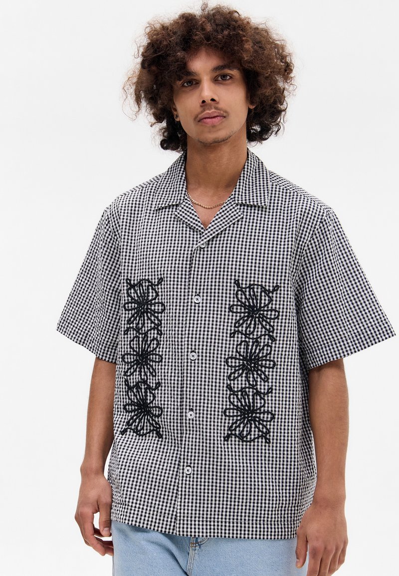 Camisa de manga curta com padrão de gingham em preto e branco, apresentando designs florais bordados a preto na parte da frente, e um fechamento de botões.