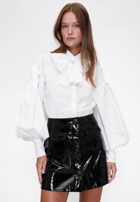 Bela srajca z velikim ovratnikom v videzu velike pentlje in napihnjenimi rokavi, kombinirana s črno sijočo mini krilom z visokim pasom, ki ima sprednje žepe in gumbe.