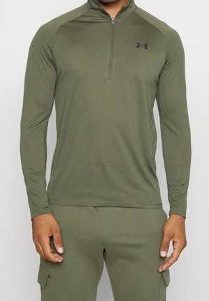 Mann trägt olivgrünes Langarm-Quarter-Zip-Oberteil und passende Hose mit seitlichen Cargotaschen, steht vor weißem Hintergrund.
