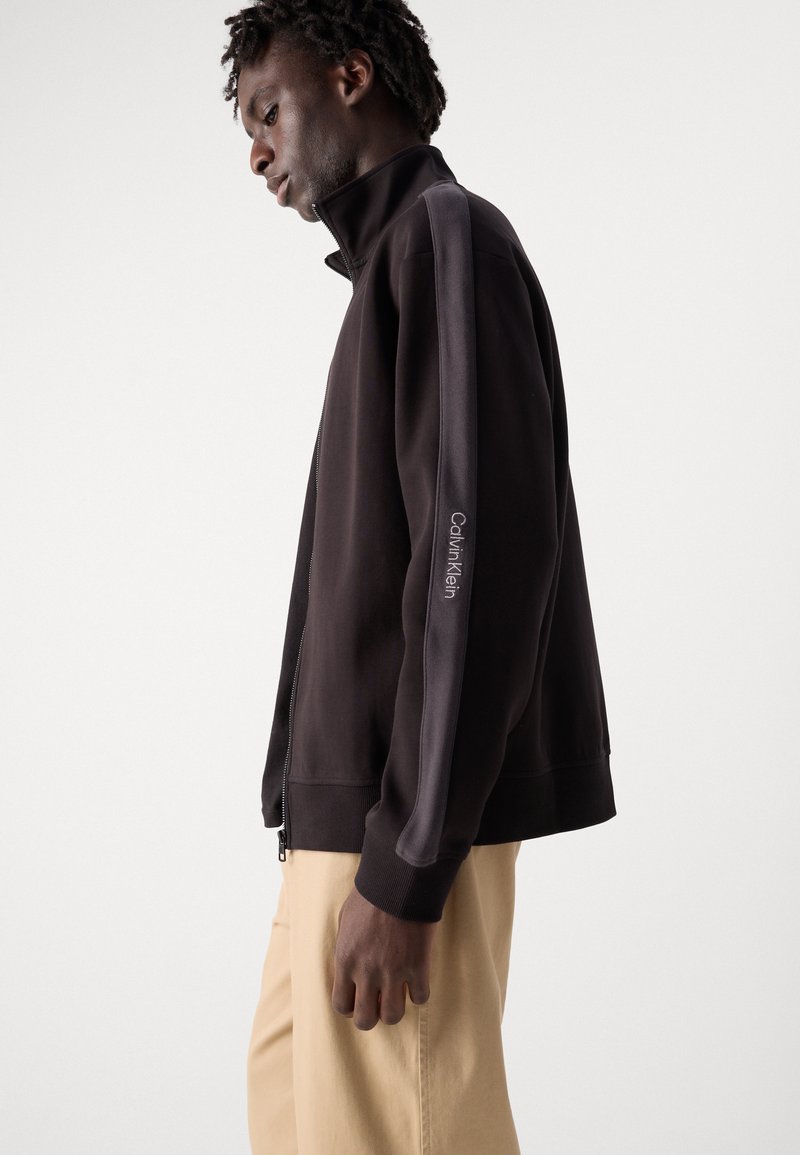 Giacca nera con zip e colletto alto, con pannelli laterali grigio scuro e logo "Calvin Klein" sulla manica. Tessuto morbido.