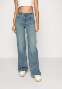 Vaqueros acampanados azul claro hechos de mezclilla, con cintura alta, bolsillos cosidos y textura desgastada, combinados con zuecos blancos.