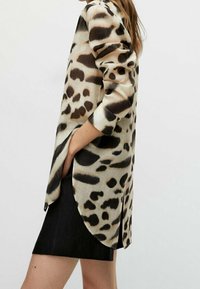 Femme portant un blouse à imprimé animal à manches longues avec un ourlet incurvé sur une jupe noire, se tenant de côté avec une main dans sa poche.