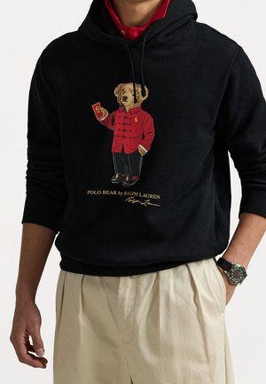 Schwarzer Hoodie mit einem Cartoon-Bären in rotem Mantel, der eine rote Karte hält. Goldener Text darunter: "POLO BEAR by RALPH LAUREN".