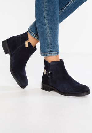 Person wearing dark blue suede ankle boots with gold zipper and buckle, paired with rolled-up blue jeans, standing on a white floor.