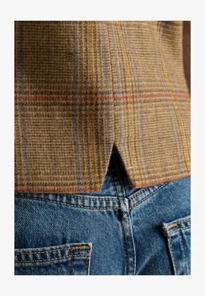 Brown plaid wool fabric with red and blue lines features a vented hem, shown over blue denim jeans with yellow stitching.