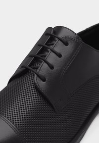 Zapato de vestir de cuero negro con textura perforada, cordones cerrados y detalle de puntera lisa mostrado en primer plano.