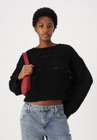 Svart heklet genser med løs, cropped design og brede ermer, kombinert med lyseblå høyhalsede jeans og en liten rød veske.