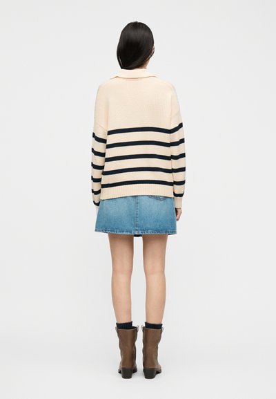 Γυναίκα που φοράει κρεμ πουλόβερ με μαύρες ρίγες, μίνι τζιν φούστα, μαύρες κάλτσες και καφέ μποτάκια στον αστράγαλο, στεκόμενη με την πλάτη προς τον θεατή σε απλό φόντο.