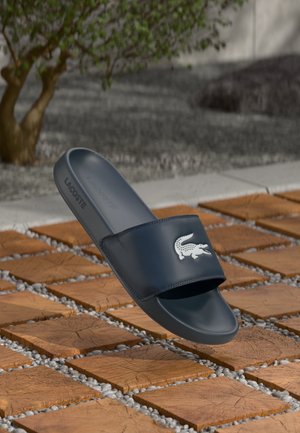 Navy blue Lacoste slide sandal with a wide strap featuring a white crocodile logo, smooth texture, and a contoured footbed.