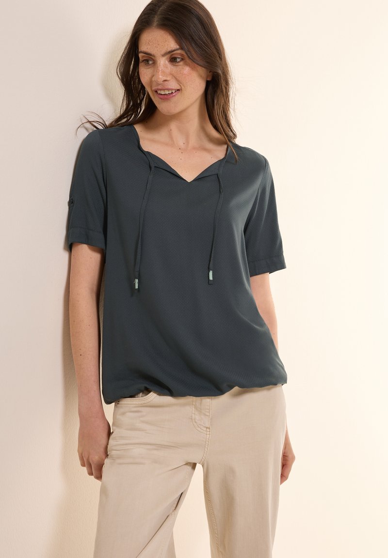 Maglietta corto maniche in teal scuro con scollatura a V e cordino. Tessuto leggero con una sottile texture. Abbinata a pantaloni beige chiaro.