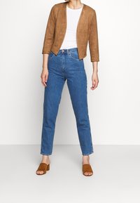 Veste courte en daim marron portée sur un haut blanc, associée à un jean bleu taille haute et des mules à talons ouvertes marron, illustrant un look décontracté.