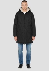 Schwarze Kapuzenparka mit Reißverschluss, kontrastierendem weichem beigem Futter, seitlichen Taschen und leicht lockerer Passform über Jeans.