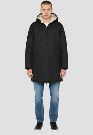 Zwarte parka met capuchon, ritssluiting, contrasterende zachte beige voering, steekzakken aan de zijkant en een iets losse pasvorm over spijkerbroek.