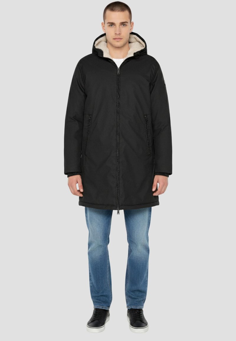 Schwarze Kapuzenparka mit Reißverschluss, kontrastierendem weichem beigem Futter, seitlichen Taschen und leicht lockerer Passform über Jeans.