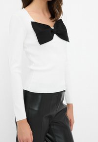 Haut blanc à manches longues orné d'un grand nœud noir au niveau du col. Associé à un pantalon en cuir noir brillant pour créer un contraste.