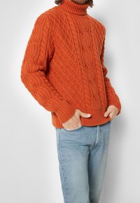 Jersey de cuello alto de punto en un vibrante color naranja, con un patrón de cables texturizados. Puños y dobladillo acanalados, combinado con jeans de color azul claro.