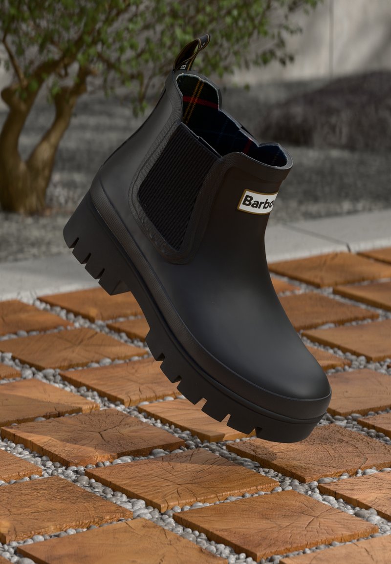 Μαύρη μπότα βροχής Barbour μέχρι τον αστράγαλο με χοντρή σόλα, ελαστικά πλαϊνά πάνελ και σχέδιο στο εσωτερικό, εκτεθειμένη πάνω από ξύλινα πλακάκια και χαλίκι σε εξωτερικό χώρο.
