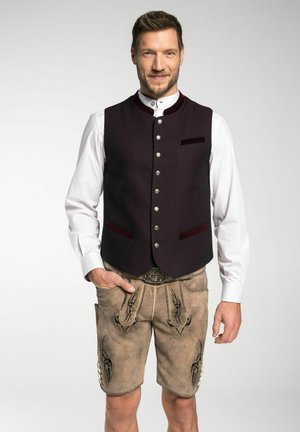 Traditionelle Tracht mit einer dunkelburgunderfarbenen Weste mit silbernen Knöpfen, ergänzt durch beigefarbene Lederhosen mit dekorativer Naht und einem Gürtel.
