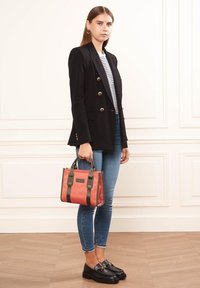 Veste noire à double boutonnage sur une chemise rayée, jeans skinny bleus, portant un sac fourre-tout corail avec des poignées vert foncé, et mocassins noirs.