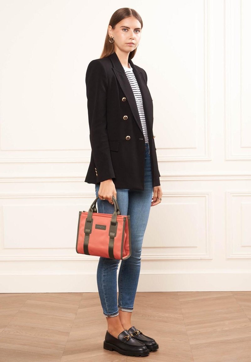 Veste noire à double boutonnage sur une chemise rayée, jeans skinny bleus, portant un sac fourre-tout corail avec des poignées vert foncé, et mocassins noirs.