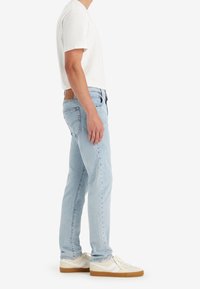 Mies, joka on pukeutunut vaaleansinisiin farkkuihin, valkoiseen lyhythihaiseen paitaan ja valkoisiin kumipohjallisiin tennareihin, seisoo profiilissa valkoista taustaa vasten.