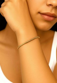 Bracciale in metallo color oro con un design liscio e sottile, appoggiato su un braccio abbronzato. Lo sfondo è scuro, creando contrasto.