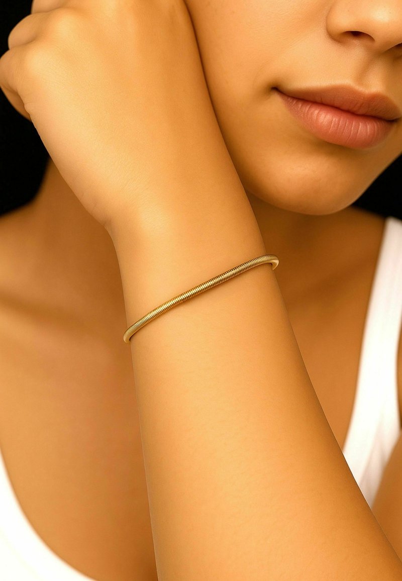 Bracciale in metallo color oro con un design liscio e sottile, appoggiato su un braccio abbronzato. Lo sfondo è scuro, creando contrasto.