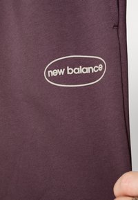 Vínová látka s hladkým povrchem, která má bílé oválné logo s nápisem "new balance" umístěné v dolním levém rohu.