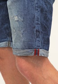 Shorts in denim blu chiaro e scuro con dettagli sbiaditi. Presentano un orlo risvoltato con accenti di cucitura rossa. Il tessuto ha una texture sbiadita.