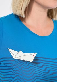 Blaues T-Shirt mit rundem Ausschnitt, verziert mit einem weißen Origami-Boot, das mit einem roten Herz geschmückt ist, und einer wellenförmigen blauen Musterunterlage.