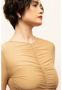 Krótki, dopasowany top w kolorze beżowym z marszczoną strukturą z przodu i z tyłu, z okrągłym dekoltem i długimi rękawami. Widać srebrne kolczyki.