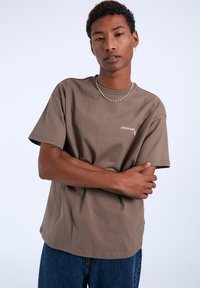 Camiseta de algodón marrón con mangas cortas y un ajuste estructurado, que presenta un pequeño logo blanco en el frente. Combinada con jeans de mezclilla azules.