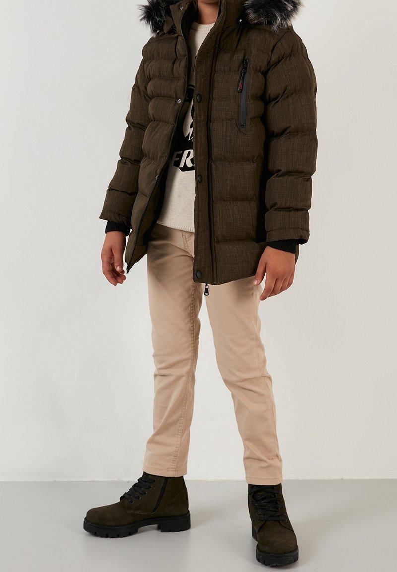 Enfant portant un manteau matelassé marron à capuche bordée de fourrure, un sweat-shirt imprimé, un pantalon beige et des bottes foncées à lacets, debout devant un fond uni.