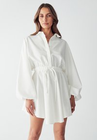 Abito camicia bianco a maniche lunghe con colletto, vita arricciata con cordoncino e fondo fluido. Tessuto morbido e leggero.