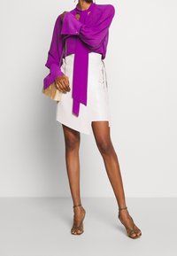 Lila Langarmbluse mit Bindebändern am Hals, kombiniert mit einem weißen asymmetrischen Rock. Model trägt grüne Riemchensandalen und hält eine Clutch.