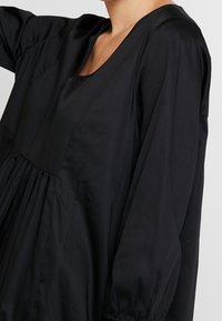 Schwarz Kleid aus glattem Stoff, mit weitem Ausschnitt, langen Ärmeln und gerafften Details an der Taille.
