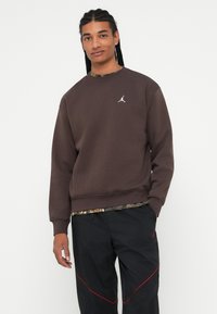 Joven con cabello trenzado que lleva una sudadera marrón con un logo blanco y pantalones negros con detalles rojos, de pie contra un fondo blanco.